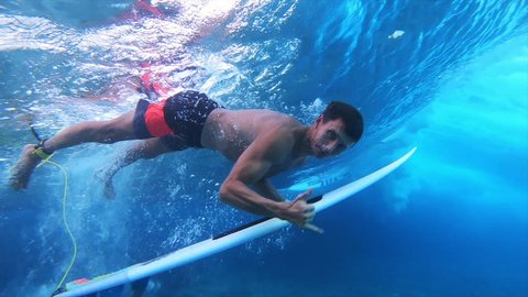 Surfer dives under the wave with the surfboard (the trick named in surfing as Duck Dive) looks into the camera and shows the Shaka sign