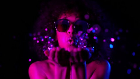 Young girl with curly hair blowing confetti on dark background in neon light. Woman celebrating, depicts joy and happiness. Success, victory, holiday, new year concept.