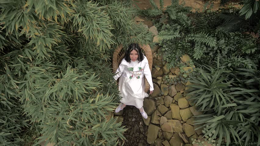 brunette in white clothes is sitting on a wicker chair on the stones, surrounded by tropical plants. The camera is directed from top to bottom.White strands in hair