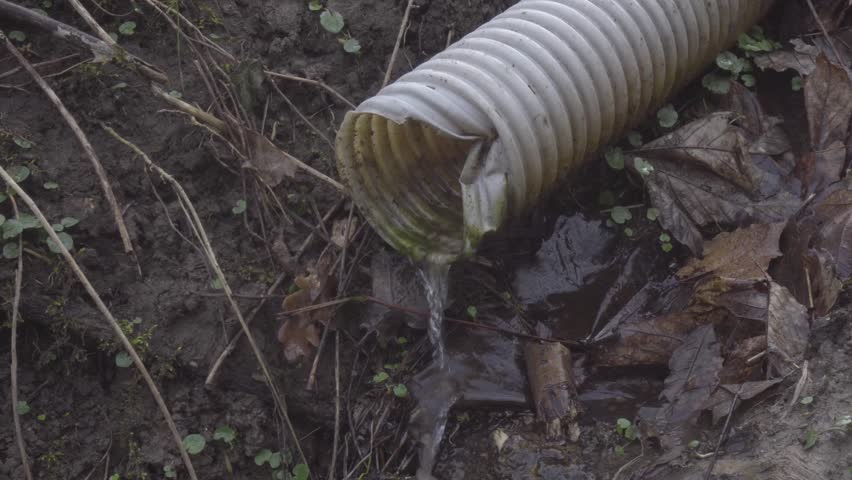 locked close shot of water leaking out of rippled plastic drainage pipe over wet earthy forest garden ground with old foilage, daylight early spring situation
