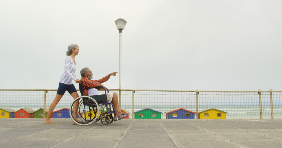 Side view of active senior African American woman pushing disabled man in wheelchair on promenade. Senior man pointing and showing her 4k