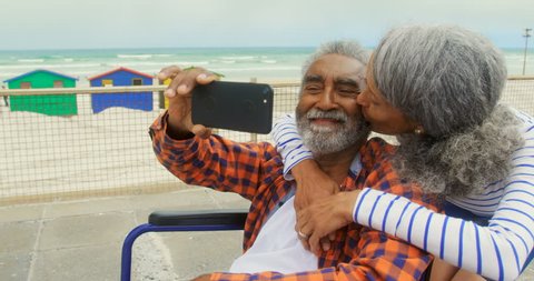 Front view of active senior African American couple taking selfie with mobile phone on promenade. Senior woman embracing and kissing him on cheek.