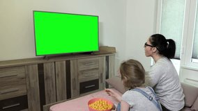 Daughter and mother eating crisps from bowl and using remote control near TV with green screen