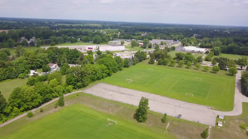 Aerial drone footage of a green park during a sunny summer day in Michigan. Baseball and soccer fields, playground, forest and trails visible.