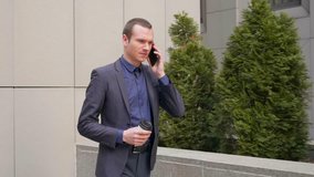 A young businessman walking down the street and aggressively leads a discussion on the phone call. The guy in a suit with a glass of coffee in his hand shows indignation by talking on a smartphone. 