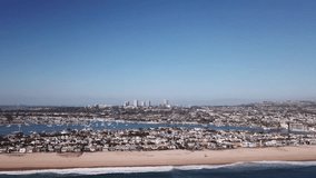 Aerial drone footage of a California beach during a beautiful and sunny summer day. Waves crashing upon the sandy shore and skyscrapers and boats visible in the background.