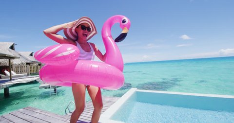 Funny vacation woman in bikini with inflatable pink flamingo pool toy mattress by swimming pool. Girl laughing having fun sunbathing enjoying travel holidays at resort luxury overwater bungalow travel