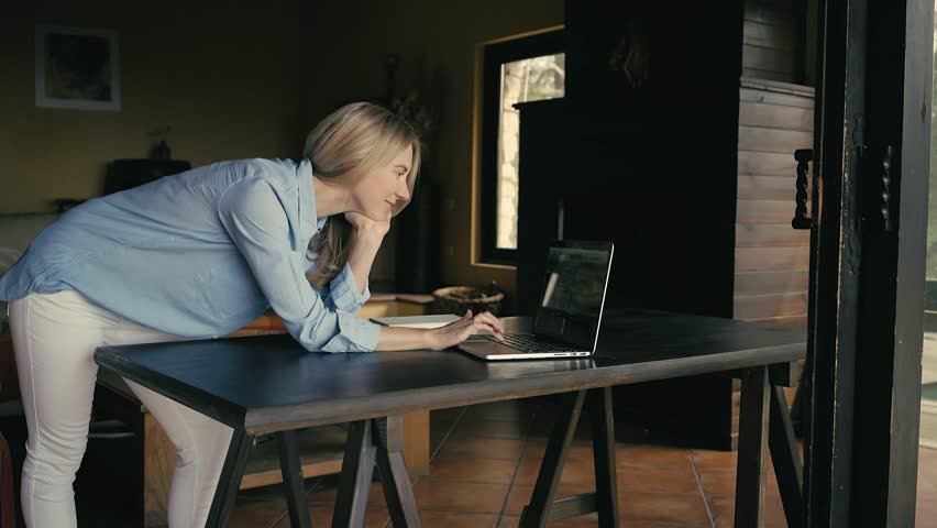 Business woman using laptop, works at home, workplace. Female drinking coffee in sunny morning near big window, Blonde woman with cup of hot tea.
