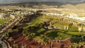 Aerial view of golf course. Cinematic 4K footage with beautiful tropical landscape, mountains, road, city modern villas. Sunset shining on golf resort. Green grass, ponds, people near the golf cart   