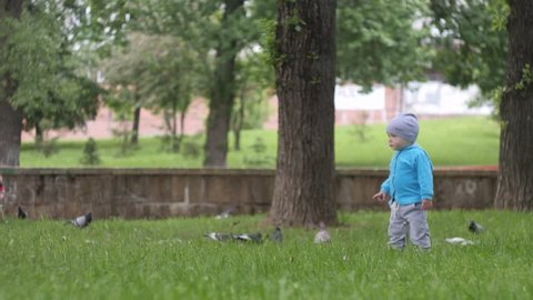 little handsome boy in blue jacket runs across the lawn to the birds in the city park with a pond on the background in slow motion 4K video
