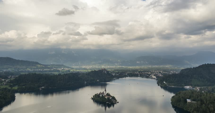 Elevated high time lapse of Bled lake with boats moving and it's island and a church on it. Clouds can be seen moving and forming above it with alps and hills in the background.