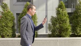 A young businessman walking down the street and happily communicates on the video call. A cheerful guy in a classic suit is smiling while talking at the video conference. 4K footage.