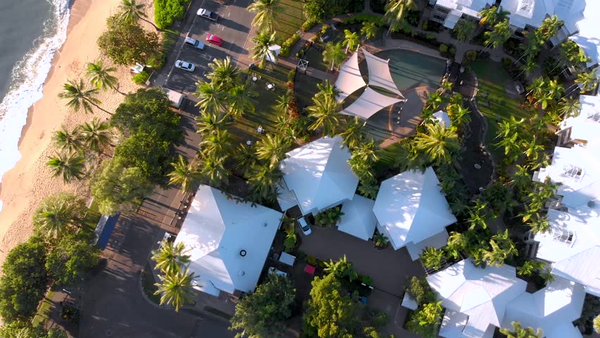 A fly up with a slow rotate and camera looking downwards above hotels and resorts on a coastline in Cairns, Australia at Sunrise with Trinity Beach across the road.
