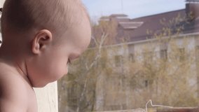 Close up of the little baby boy is sitting by the window, look into it, then into the camera and point to something outside the window on the background of a tree behind the window in slow motion 4K
