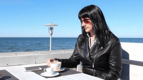 young brunette woman in black leather jacket and sunglasses drinking coffee in an outdoor cafe near the sea on sunny day