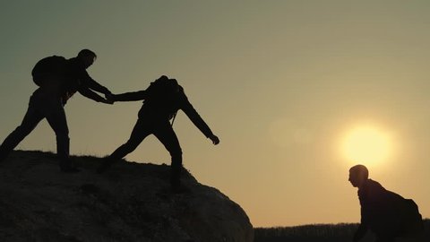 Silhouettes of tourists climbing mountain cliffs against the backdrop of a sunset, helping each other's hand. Help in the mountains and teamwork in hiking. Teamwork concept.
