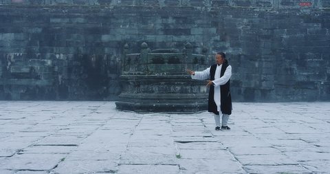 An Asian master of tai chi martial arts demonstrates tai chi in winter with snow falling in Wudang mountain monastery China. Slow motion, red cinema camera hand held. 
