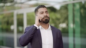 Serious businessman talking by smartphone on street. Slider shot of focused bearded man in suit jacket standing outdoor and having phone conversation. Communication concept