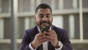 Smiling man using cell phone outdoor. Handsome bearded businessman using smartphone and laughing, handheld shot. Technology concept