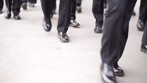 Many men in identical black patent leather shoes and black trousers walk down the street.