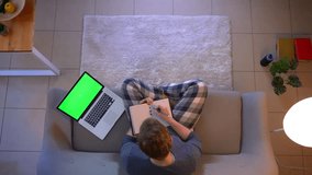 Closeup top shoot of young businessman working and taking notes using the laptop with green screen while sitting on the sofa indoors at cozy home