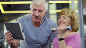Medium shot of Caucasian elderly man in sportswear standing near his wife doing exercises with dumbbell and showing her video lesson on tablet