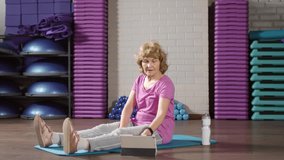 Wide shot of aged Caucasian woman sitting on fitness mat, turning on video lesson on tablet and starting pumping arms muscles using dumbbell