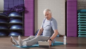 Wide shot of aged Caucasian man sitting on fitness mat, turning on video lesson on tablet and starting doing exercises using dumbbells