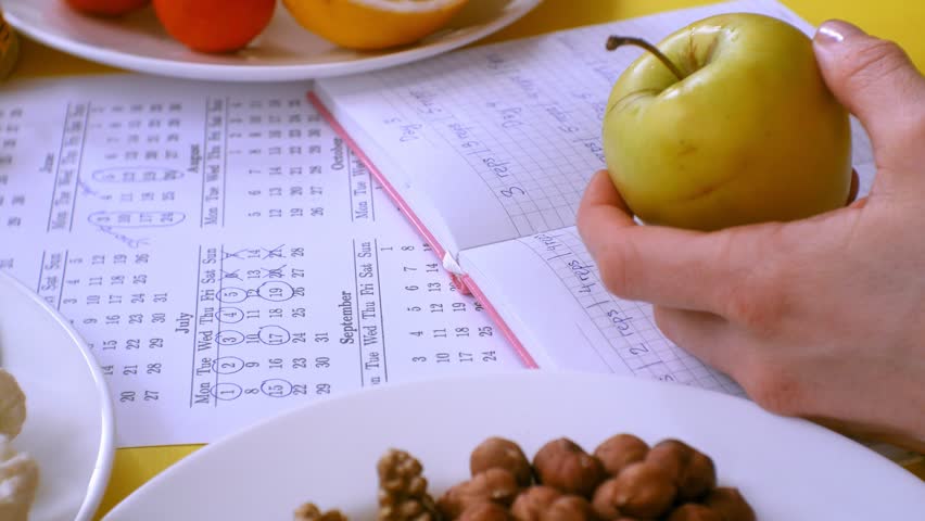 A woman on a yellow background makes notes in a sports notebook, in one hand holds a green apple, looks at the calendar, there is a measuring tape and plates with nuts, fruits, vegetables on the table