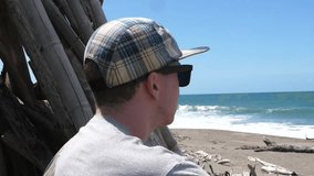 Young male in a cap and sunglasses sitting by a wooden tipi looking out to sea in Fiji