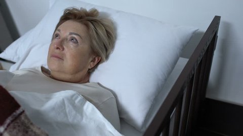 Volunteer wiping tears of elderly terminally ill female patient lying in sickbed