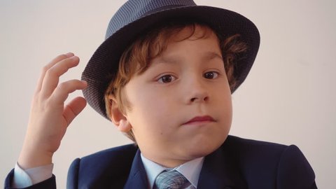 Portrait of child boy looks like a businessman in hat and suit in his office is looking through magnifier at light background, front view.