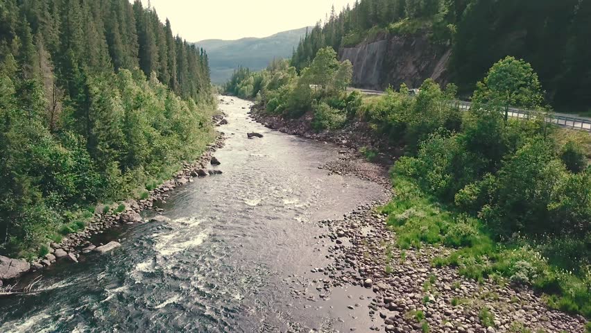 Close and slower forward drone shot over a river next to a road in a forest. Mountains are shown in the bachground. 4k quality video in Norway