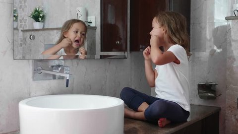 Little cute girl brushes her teeth with dental floss while sits on the table near the sink at domestic bathroom in front of the mirror.