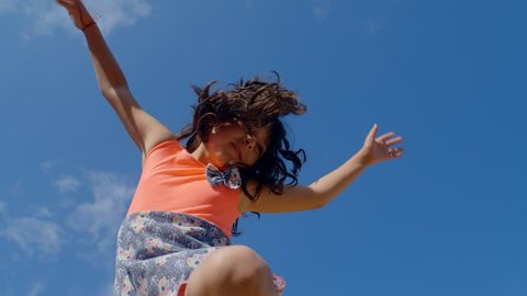 A cute 9-10 years old girl with curly hair having fun jumping at trampoline blue sky background, 4k slow motion low angle view