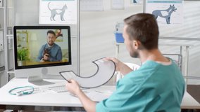 Medium shot of male vet in scrubs sitting at his desk and consulting man with cat via video call. He is talking and showing how to put on e-collar