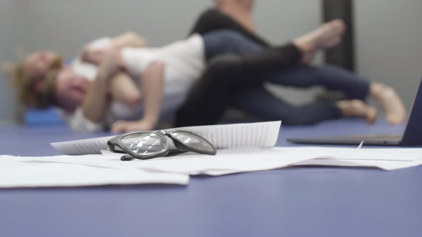 Two men in white shirts wrestling and doing grips lying on the floor in the background. Papers, laptop and glasses on the floor in the foreground