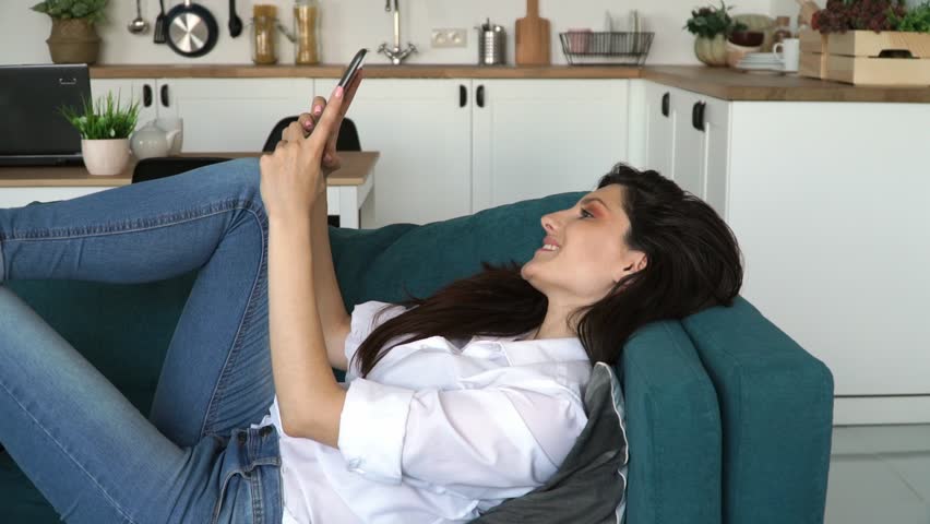 Young attractive smiling caucasian woman with long black hair uses tablet while lies on sofa at home. Girl typing on tablet and talks to friends.
