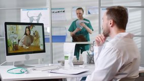 Medium shot of male vet in white coat sitting at his desk and talking on video call with female dog owner