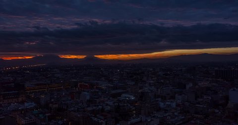 Aerial hyperlapse of colorful sunrise in historical center with the Zocalo main square the Mexican flag and the volcanoes at the back.