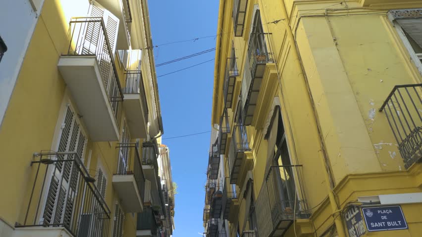 Camera tilts down from the tops of two yellow buildings to street level, with a shot down the narrow street between the buildings.