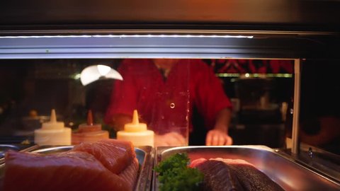 Chef preparing Sushi roll behind salmon fish glass container in a cafe, sliding shot