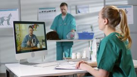 Medium shot of female veterinarian in scrubs sitting at her desk and having video call with man with sick cat. She is nodding and making notes in medical form