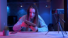 Closeup shoot of young attractive female blogger with dreadlocks playing video games on the tablet getting emotional streaming live live with the neon background indoors