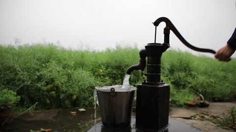 hand pump in front of a countryside village house, providing groundwater or underground water from aquifer as drinking water supply for villagers. Rapeseed weeds field in background