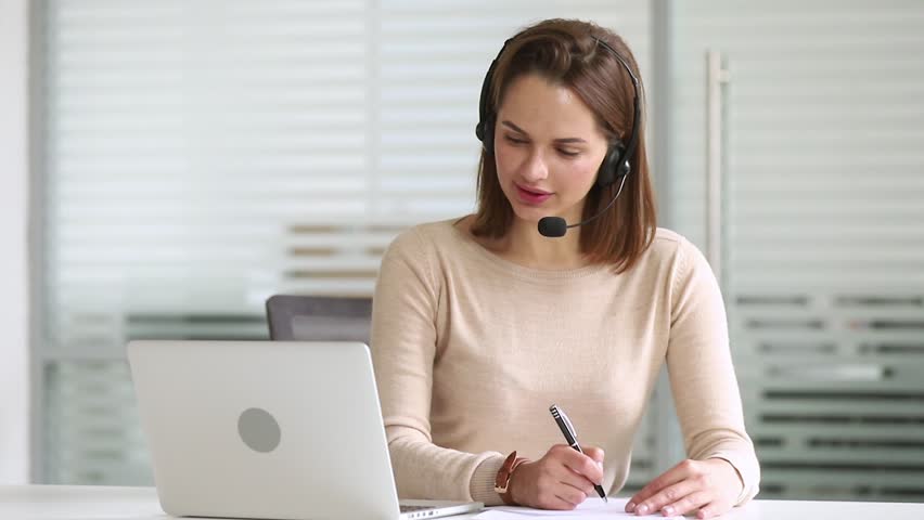 Business woman customer support service agent receptionist or interpreter wearing headset looking at laptop make notes listen translate talk by conference call on consult client on video chat Royalty-Free Stock Footage #1028495384