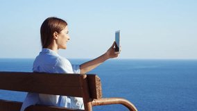 Young woman sitting relaxing on bench at edge of cliff talking via messenger app having video chat on cellphone. Female enjoying breathtaking view of blue Mediterranean sea using mobile phone