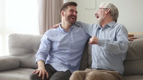 Happy generations old father laughing embracing young son giving fist bump sit on sofa at home, friendly senior dad hug adult man talking joking having fun enjoy time together and good relationships