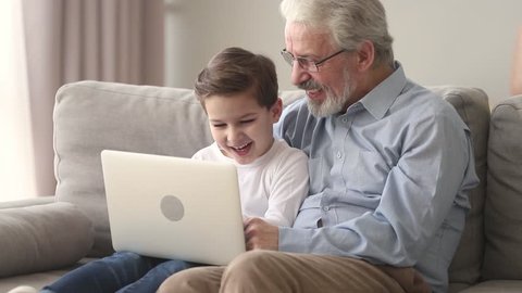 Happy old grandfather and little cute grandson laughing looking at laptop sitting on sofa, grandkid small boy sit on senior grandpa lap talking teaching using laptop having fun with computer at home