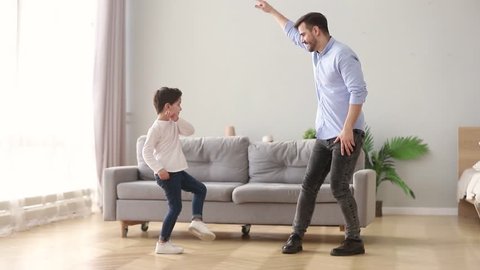 Happy young dad and cute little kid son dancing having fun in living room together, funny active small boy laughing imitating father moving to music, daddy child playing at home on weekend leisure